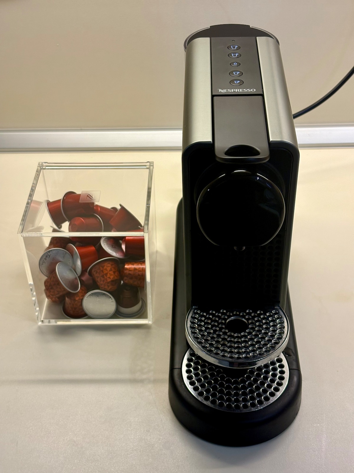 A coffee machine is positioned on a light surface, featuring a rounded water reservoir and control buttons on top. Beside it, a transparent container holds an assortment of colorful coffee capsules, showcasing various shapes and shades.