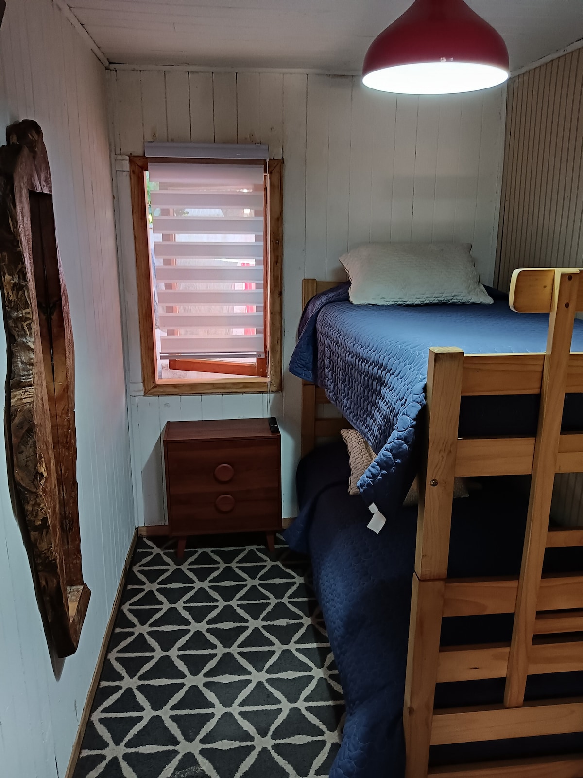 A compact sleeping area features a wooden bunk bed with navy bedding. A small nightstand with two drawers is positioned beside the bed. Natural light enters through a window adorned with blinds, while a patterned rug lines the floor.