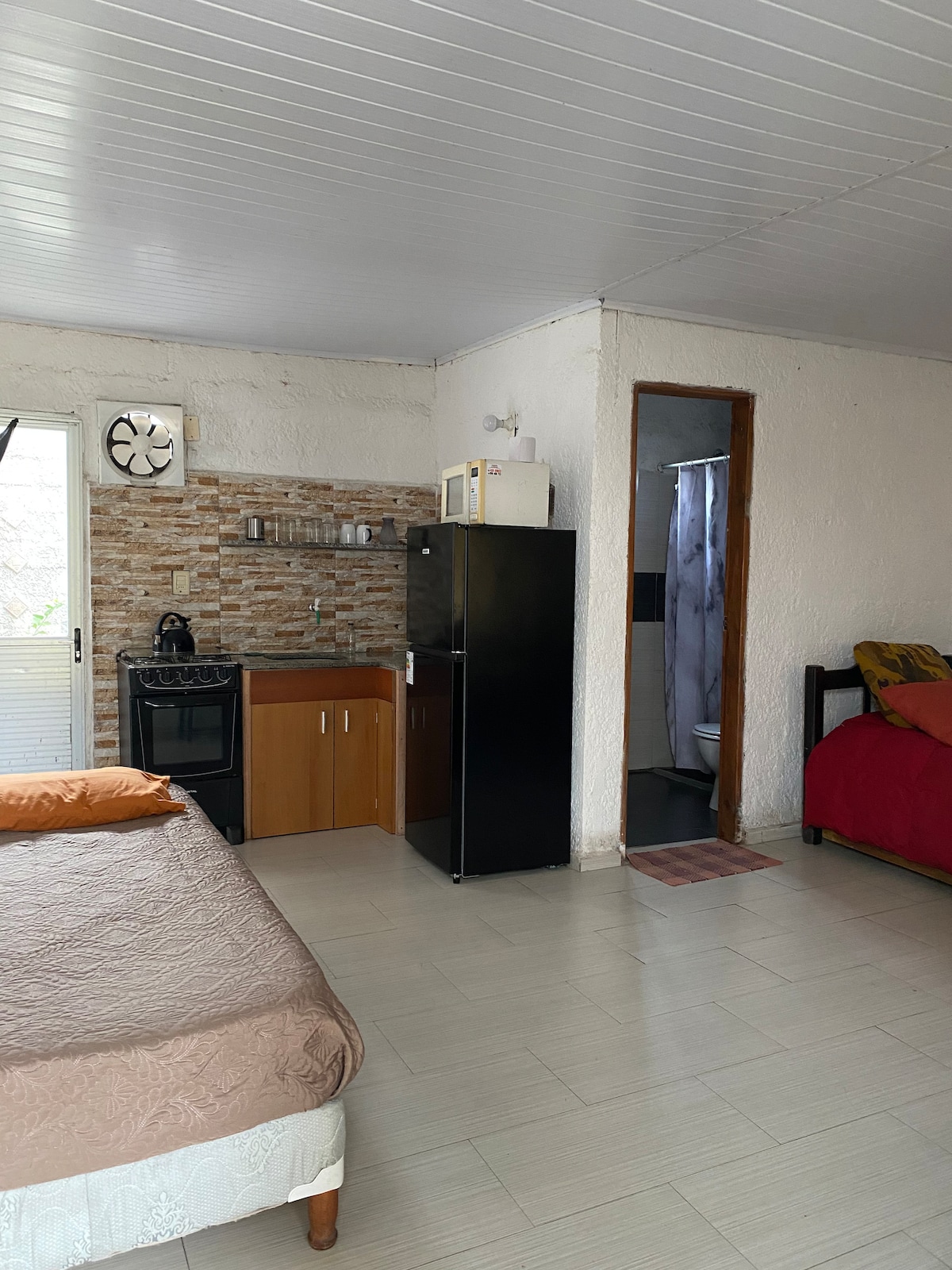 A well-organized living space features a bed with a light-colored cover and an orange accent pillow. To one side, a compact kitchenette includes a refrigerator, stove, and wooden cabinetry. The bathroom door is visible in the background, while a window provides natural light.