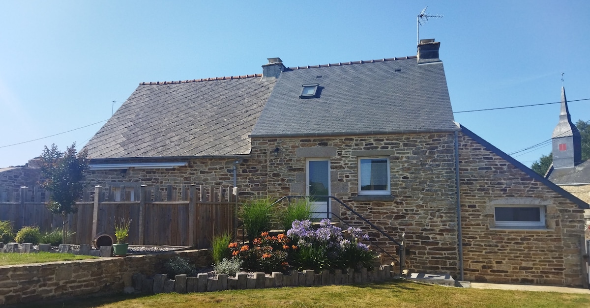 The exterior of the stone cottage features a sloping gray roof and a well-kept garden with colorful flowers. A pathway leads to the entrance, framed by lush greenery. The surrounding countryside is visible under a clear blue sky.