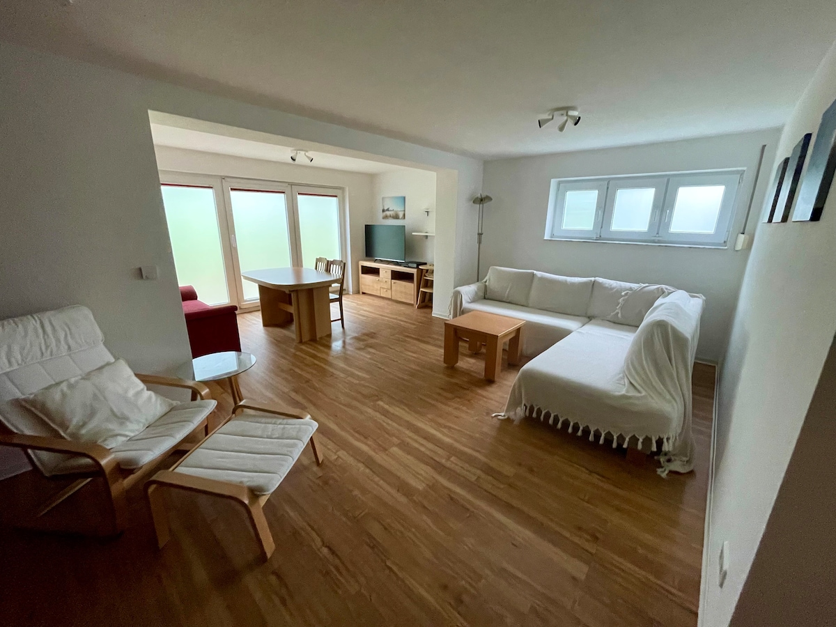 A bright living space features wooden flooring and natural light coming through large windows. A cozy white sofa is accompanied by a wooden coffee table and a sleek dining table. A comfortable armchair is present, enhancing the room's functionality.