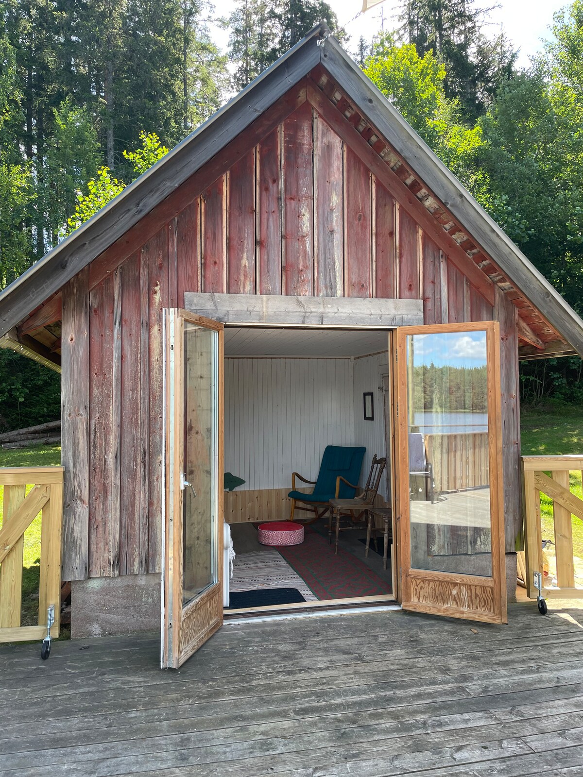 An inviting wooden cabin is presented, featuring a large double door that opens to a wooden deck. Inside, a chair and a small table create a quiet space. The red exterior of the cabin blends with the surrounding greenery, providing a serene outdoor view.