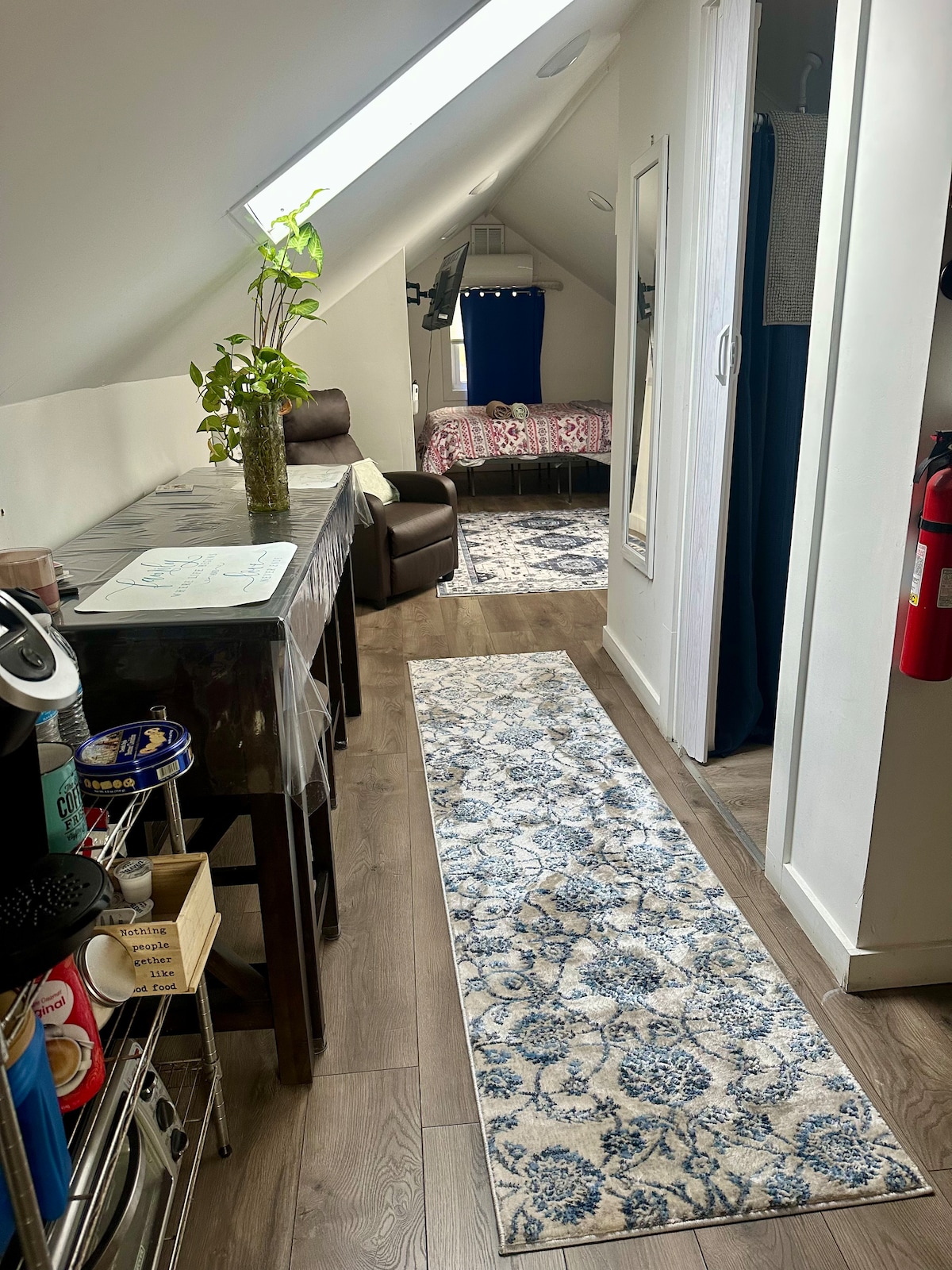 A narrow hallway leads to a cozy living space, featuring a patterned runner rug on wooden flooring. On the right, a kitchen area is visible with essential appliances, while a spacious bed is located at the far end, framed by blue curtains. A mirror reflects the light from the skylights.