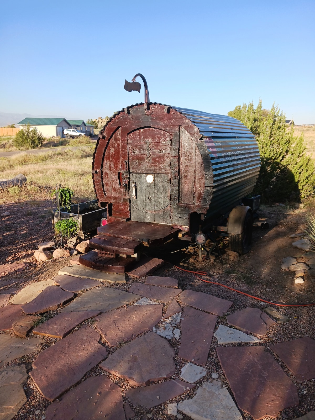 The exterior of a cozy gypsy caravan is showcased, featuring a curved metal structure with a wooden door. A stone pathway leads to the entrance, surrounded by natural landscapes and sparse greenery in the background under a clear blue sky.