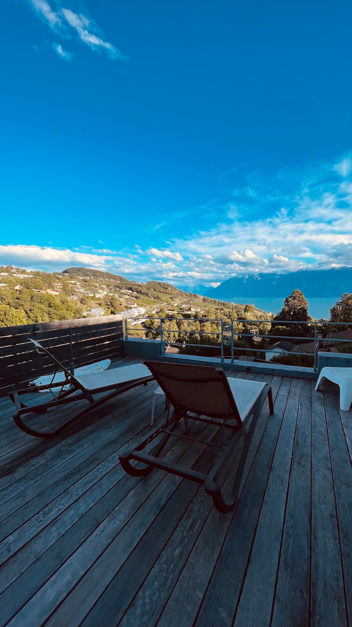 An upper terrace features sun chairs positioned on wooden decking, overlooking a scenic view of the lake and mountains. The expansive sky enhances the tranquil setting, complemented by greenery in the foreground and distant hills.