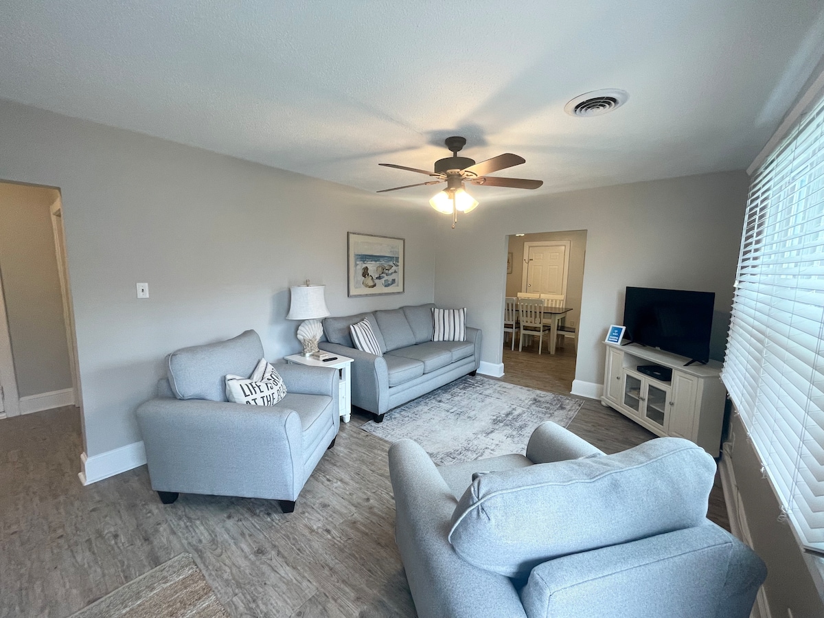 The living room features comfortable seating with two gray armchairs and a sofa, complemented by a light ceiling fan. A wall-mounted TV and side table are visible, along with a rug on the floor. Natural light filters in through a window with blinds.