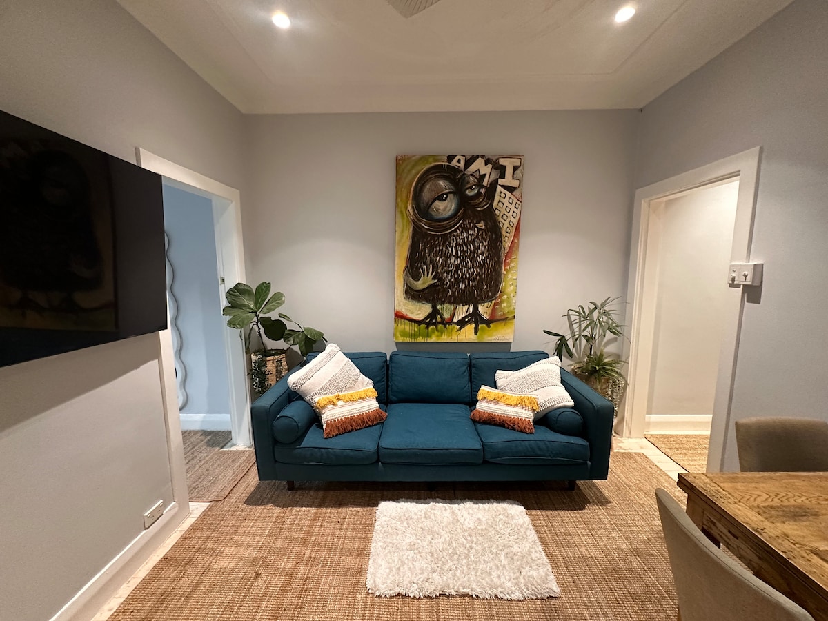 A cozy lounge area features a deep blue sofa adorned with decorative cushions. A vibrant owl painting adds a splash of color to the light grey wall. A plush area rug lies beneath, and a green plant complements the inviting space.