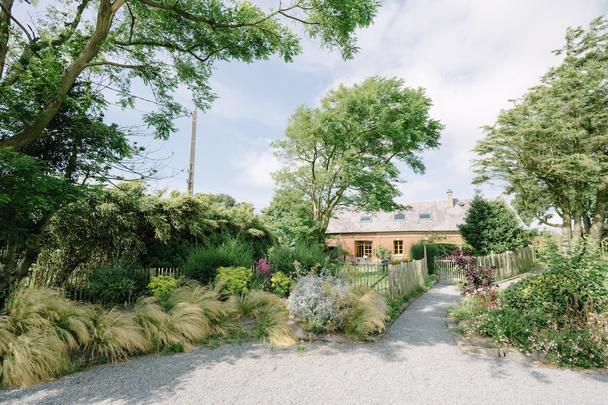 The exterior of the property is framed by lush greenery and vibrant foliage. A gravel path leads through a well-maintained garden, showcasing various plants and flowers, to the welcoming brick house under a clear sky.