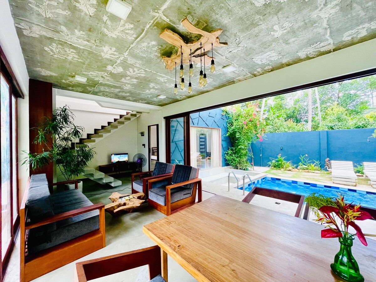 An open living area is captured, featuring a wooden table surrounded by dark-framed seating. A modern ceiling fixture provides ambient lighting. Large glass doors open to a tranquil outdoor space with a swimming pool and sun loungers, framed by greenery and colorful plants.
