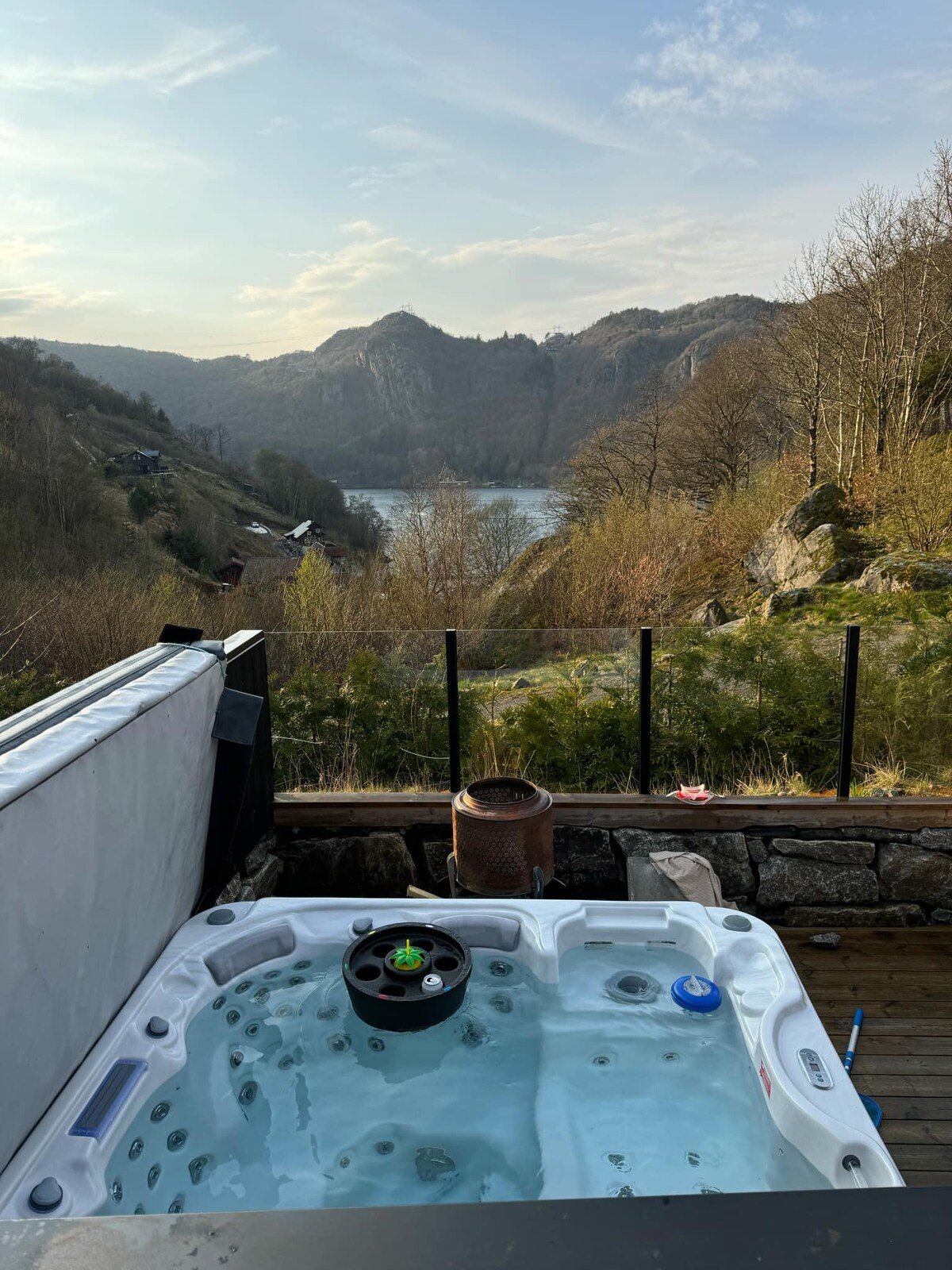 A hot tub is positioned on a deck, overlooking a scenic landscape of mountains and a tranquil body of water. Surrounding the hot tub, lush greenery contrasts with the rocky terrain, while a protective glass barrier provides unobstructed views of the natural surroundings.