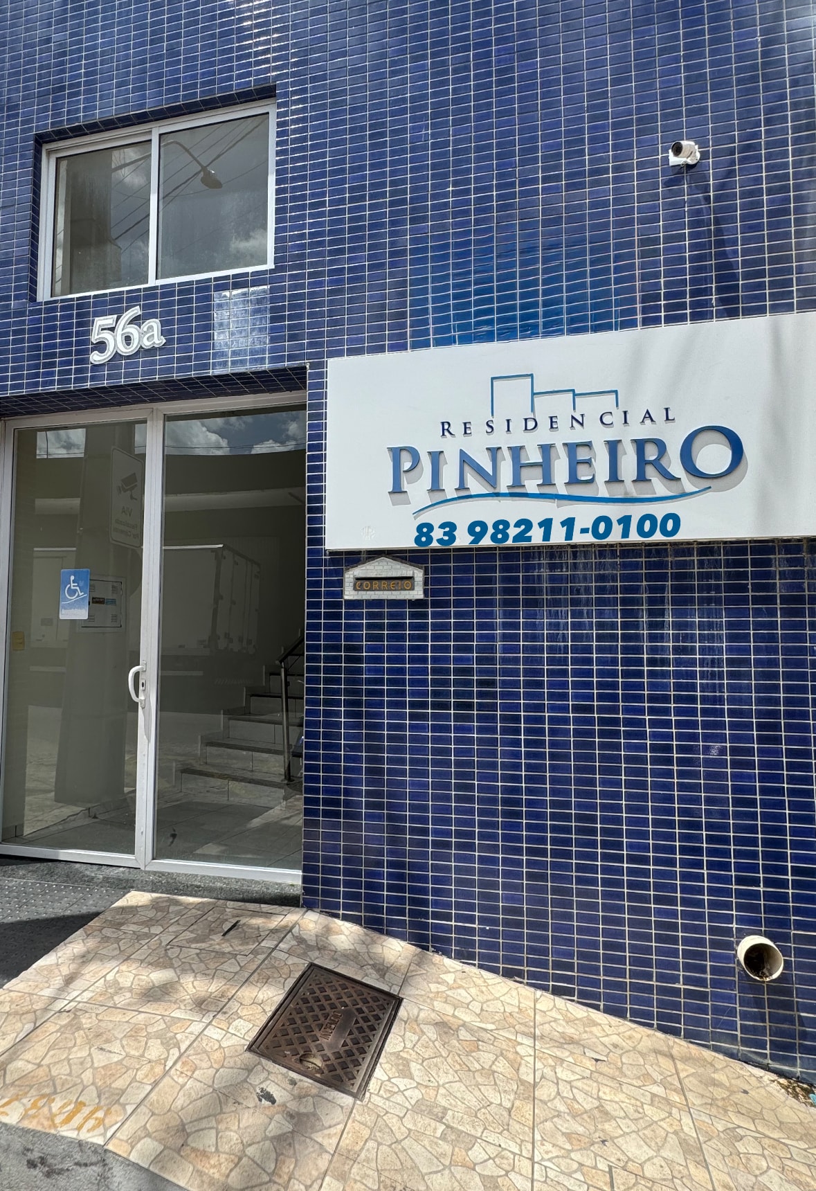 The entrance to the building is framed by glossy blue tile, displaying the name 'Residencial Pinheiro' prominently. Nearby, a glass door provides access to the interior, with a staircase visible in the background. A stone path leads to the entrance.