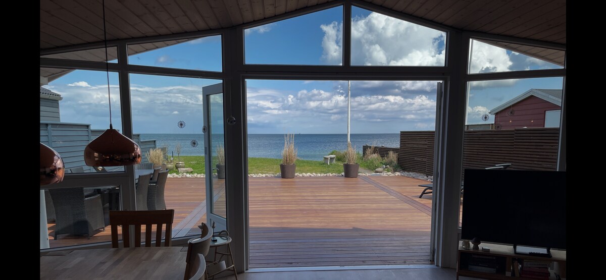 A spacious interior is highlighted by large glass doors that open to a wooden deck overlooking the beach and sea. The area features a dining table and modern seating arrangements, offering a clear view of the tranquil coastal scenery beyond.