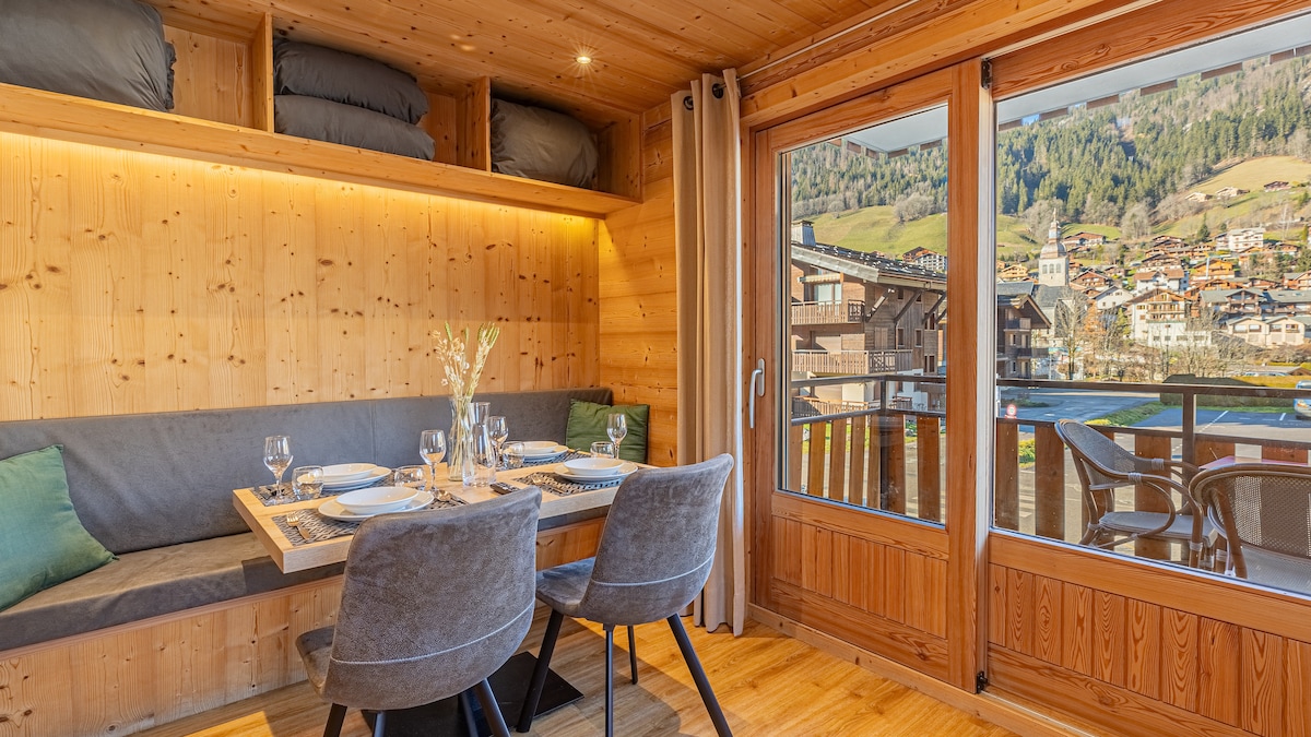 A modern dining area features a wooden table set for four, surrounded by upholstered chairs. Large windows provide a view of the mountains and village. Soft lighting highlights the wooden interior, and storage above includes neatly arranged blankets.
