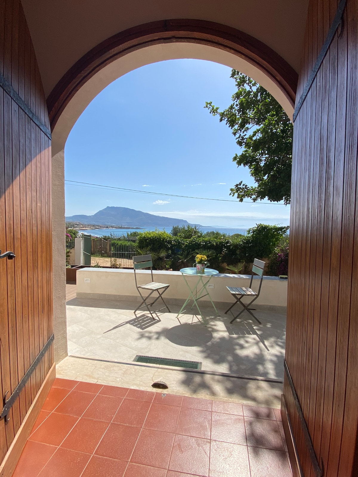 A welcoming entrance is framed by wooden doors, leading to a small patio with two metal chairs and a round table adorned with a flower vase. Scenic views of mountains and the sea extend beyond the patio, illuminated by natural light.