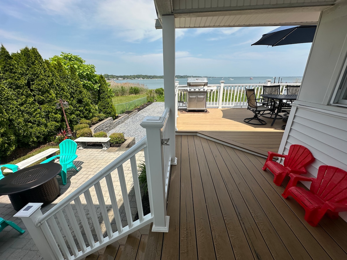The outdoor deck features a grill, dining table, and two red adirondack chairs. Green landscaping and a sandy beach are visible in the background, alongside the water. An umbrella provides shade, and the overall space is designed for relaxation and outdoor dining.