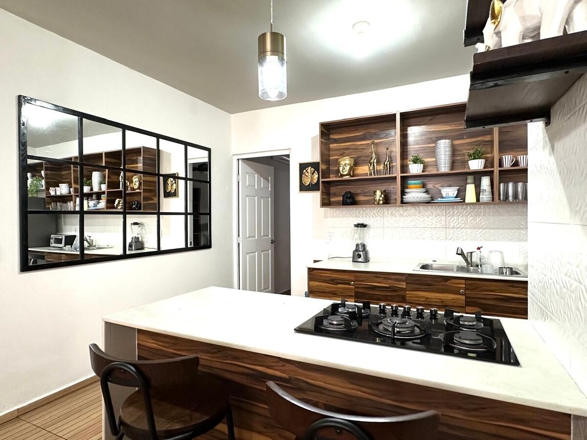 A modern kitchen features a central island with a gas stove and bar seating. Wall cabinetry displays decorative items, and a reflective wall mirror enhances the sense of space. Essential appliances, including a blender and a sink, are visible in the efficient layout.