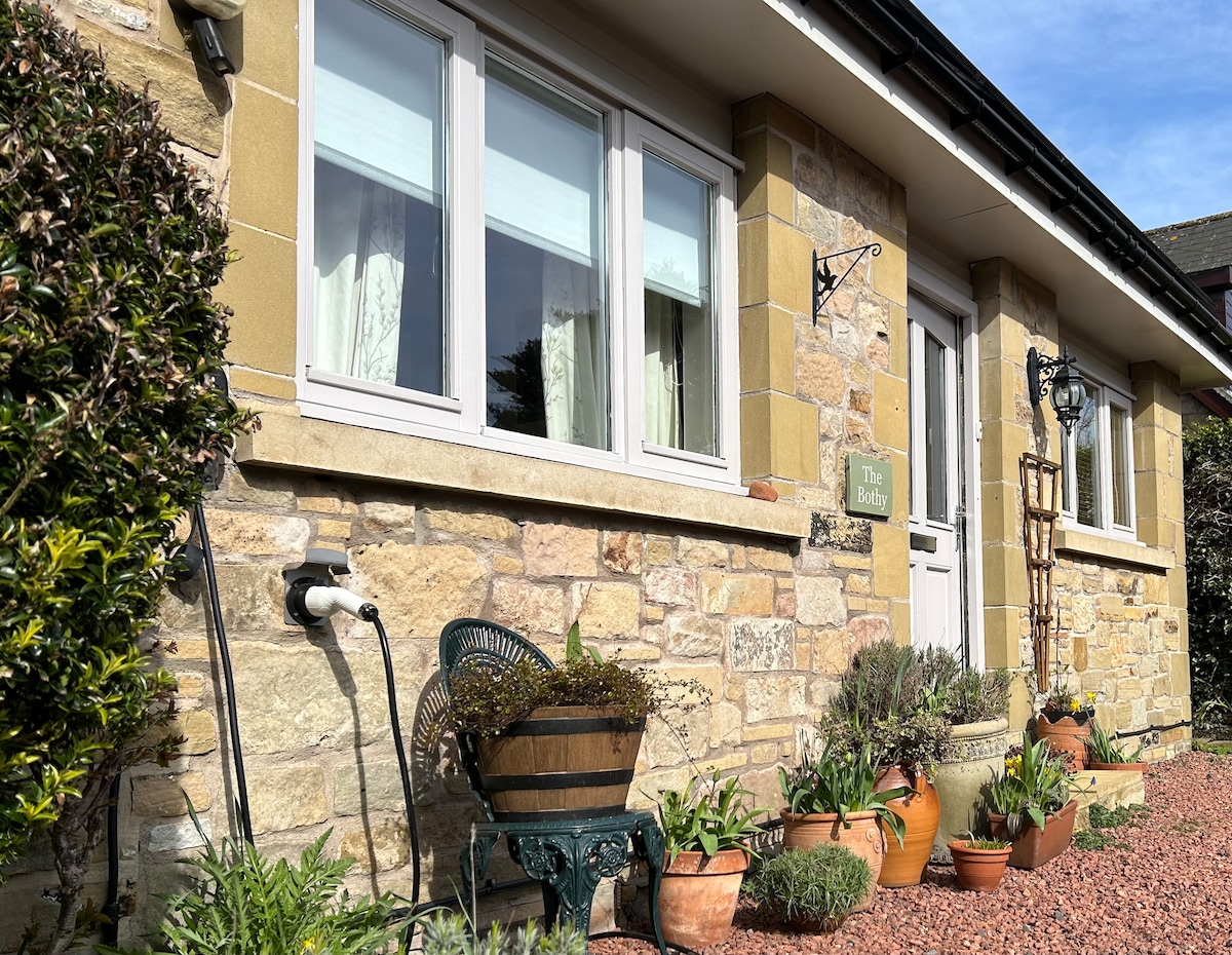 The exterior of the annex features a mix of stone and soft-colored walls, complemented by several potted plants. Large windows provide natural light, and a welcoming entrance is marked by a sign reading 'The Bothy.' The surrounding gravel area adds charm to the property.