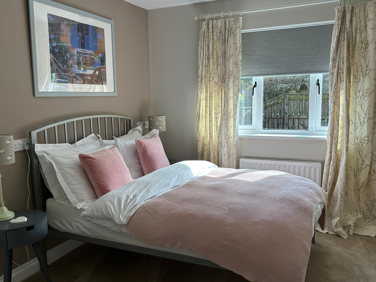 A comfortable double bedroom is presented with light-toned walls and soft furnishings. The bed is adorned with a pink throw and decorative pillows. Natural light filters through a modern window, framed by patterned curtains, providing a serene atmosphere.