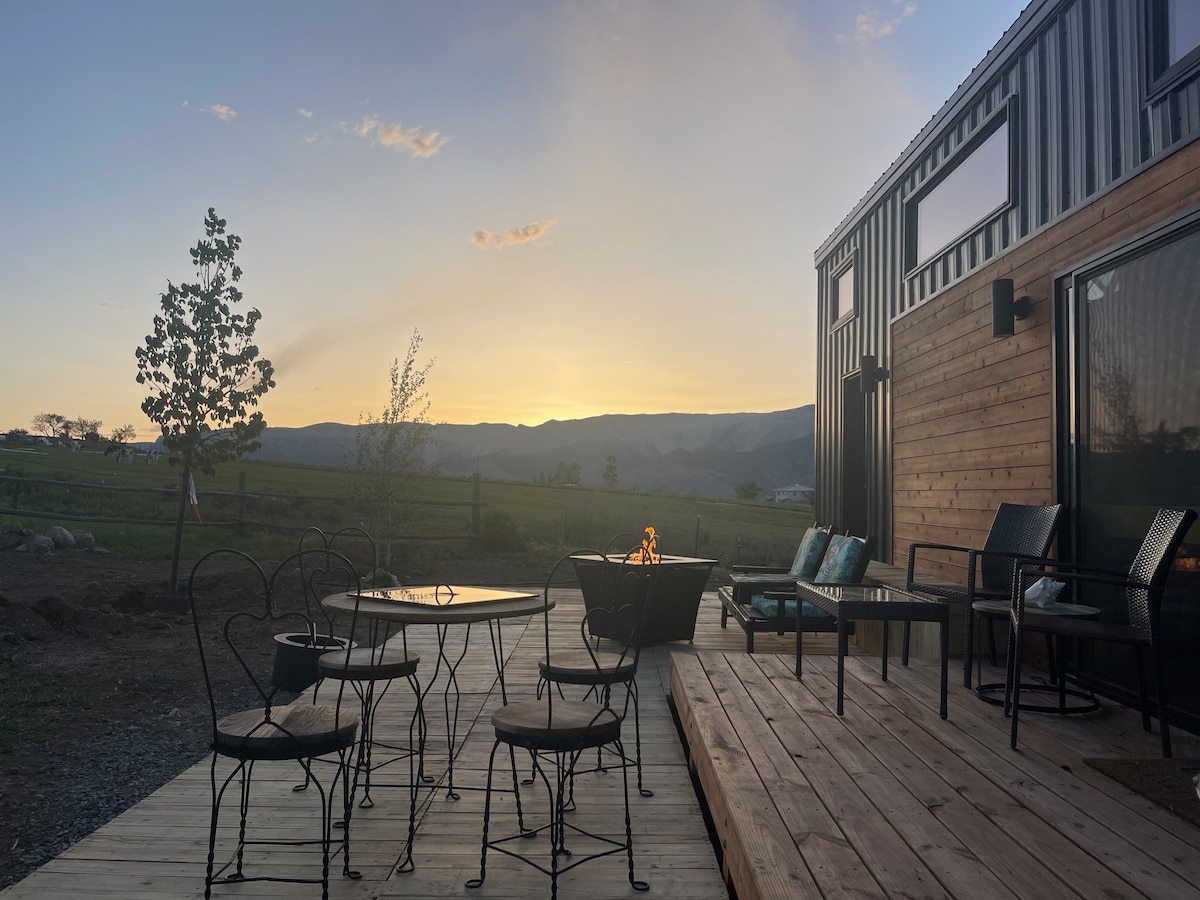 An outdoor deck is furnished with a glass-top table surrounded by four metal chairs, alongside a cozy seating area. A fire pit is visible, with mountains and a sunset in the background, creating a serene outdoor space with views of the open landscape.