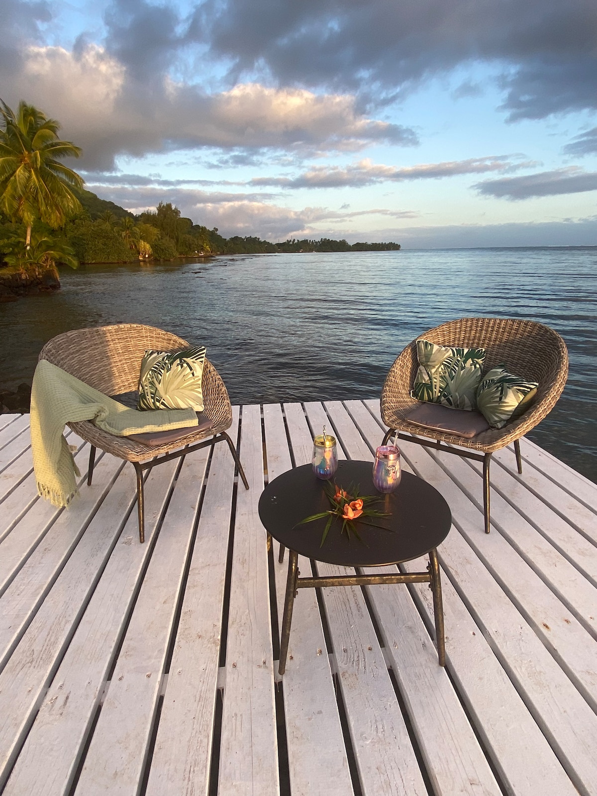 A private deck extends over calm waters, featuring two woven chairs with tropical-patterned cushions and a small black table. A light green blanket rests on one chair, while two colorful drinks and a small floral arrangement sit atop the table, framed by lush surroundings.