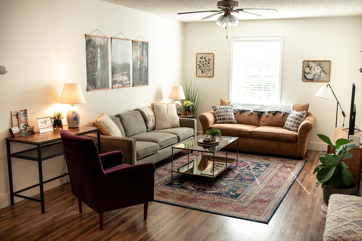 A cozy living area is presented with two sofas, one light gray and one brown, complemented by a glass coffee table resting on a patterned rug. A modern ceiling fan and soft lighting from two lamps create a warm ambiance. Wall art and plants add subtle touches of decor.