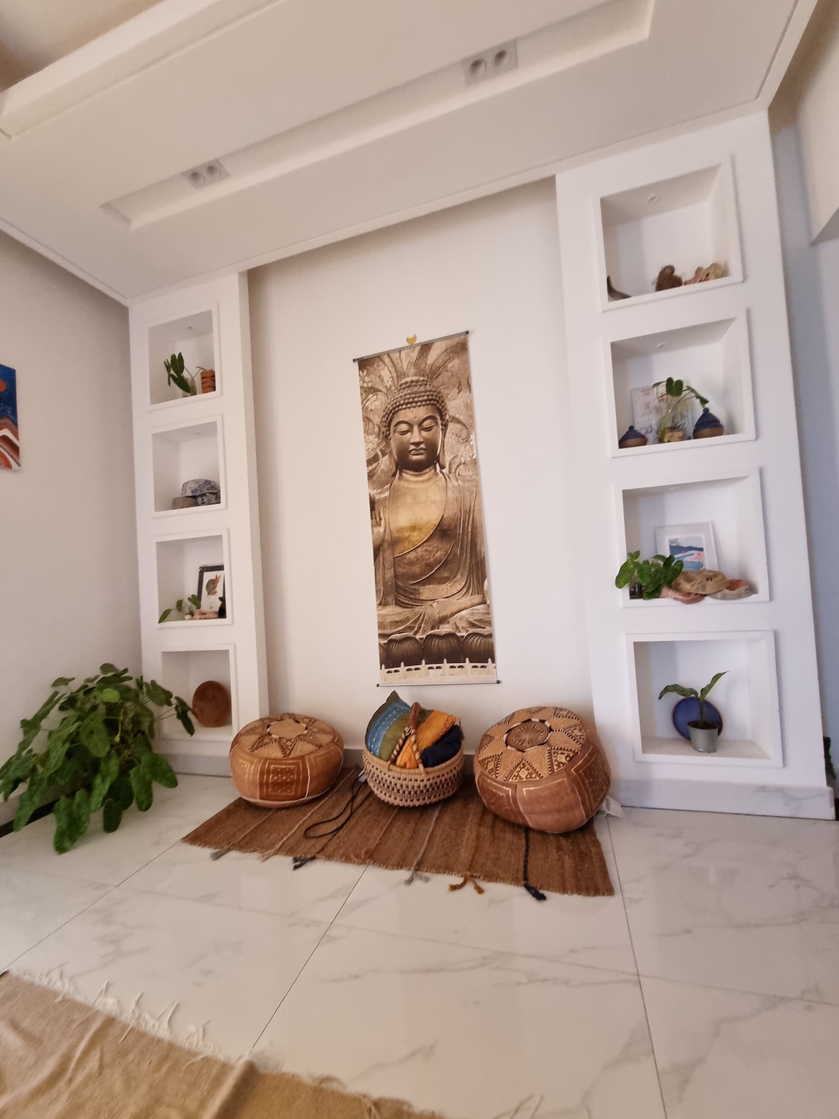 A calming interior space features white built-in shelves displaying decorative items and plants. Two intricately woven baskets sit on a textured mat, complemented by a large wall hanging that adds cultural depth to the environment.