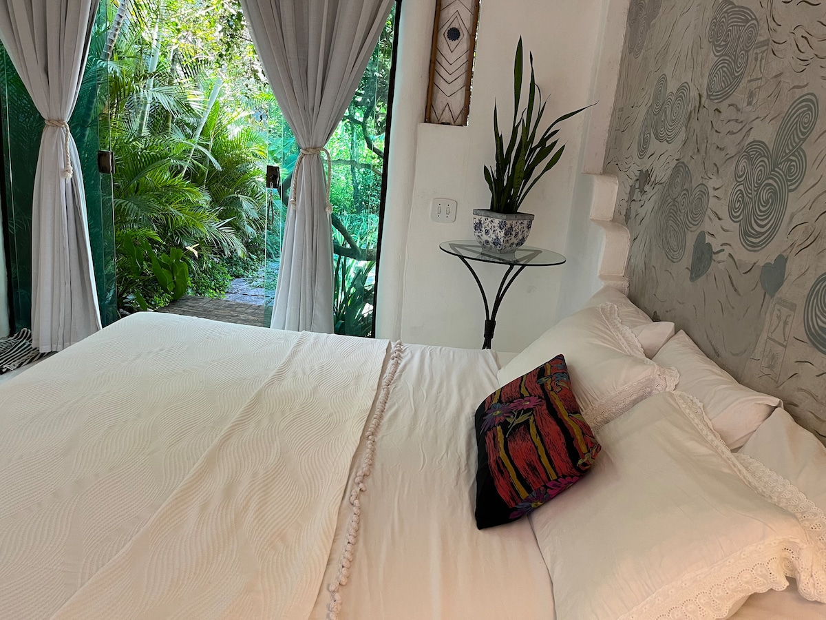 A spacious bed is positioned near large glass doors, offering a view of the lush greenery outside. Soft white linens are paired with a colorful decorative pillow. Light flows into the room, highlighting the natural textures of the walls and the decorative plant on the side table.