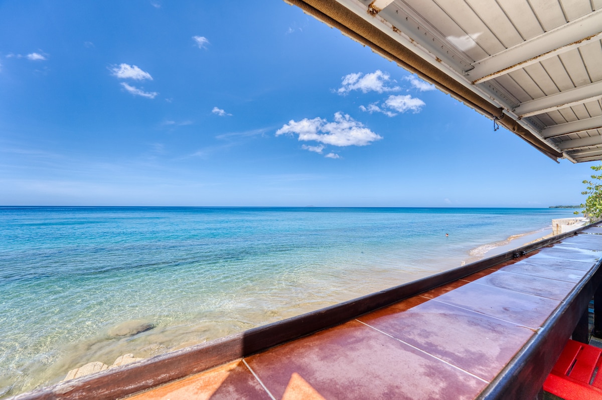 A panoramic view of the calm ocean is captured from a covered outdoor patio. The clear blue waters gently meet the sandy shoreline, while soft clouds drift above in a bright sky. A wooden bar surface is prominently displayed in the foreground.