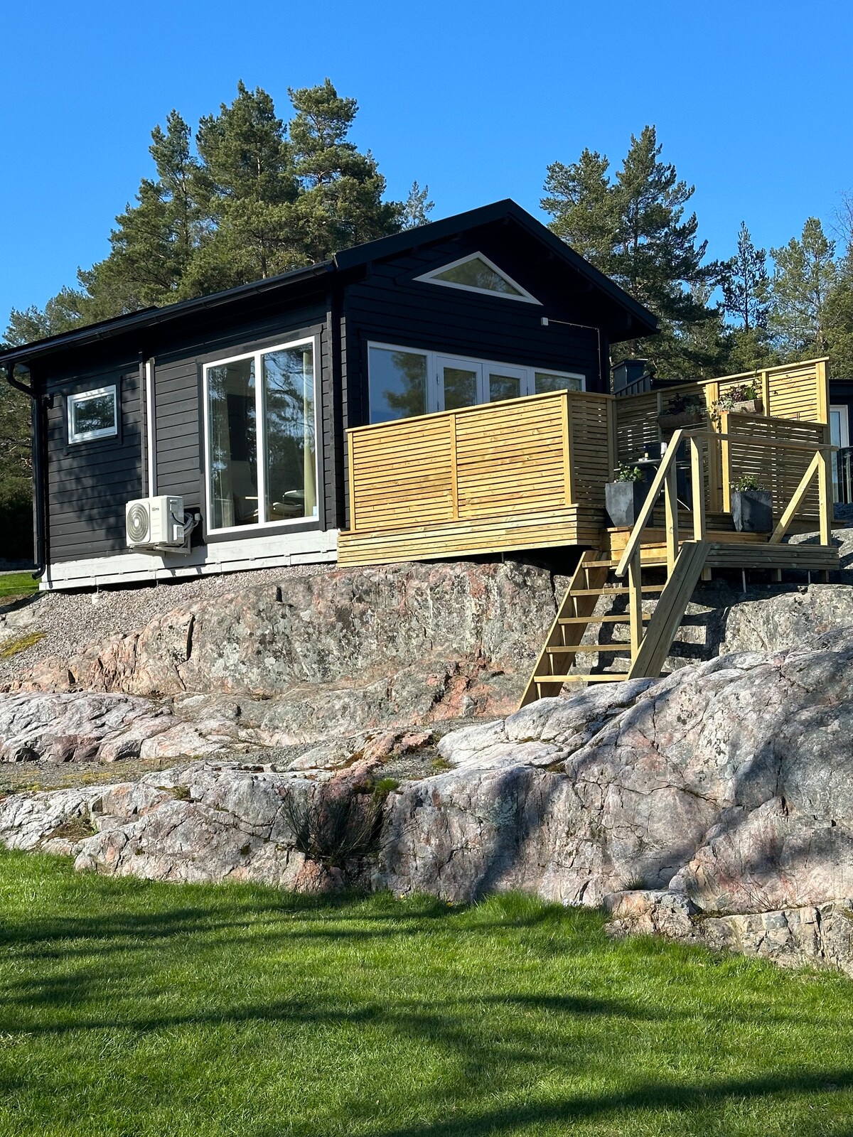 A modern, black cottage is situated on a rocky elevation, surrounded by lush greenery. Large windows offer ample natural light, while a spacious deck equipped with planters provides an inviting outdoor area. A set of stairs leads from the deck to the grass below.