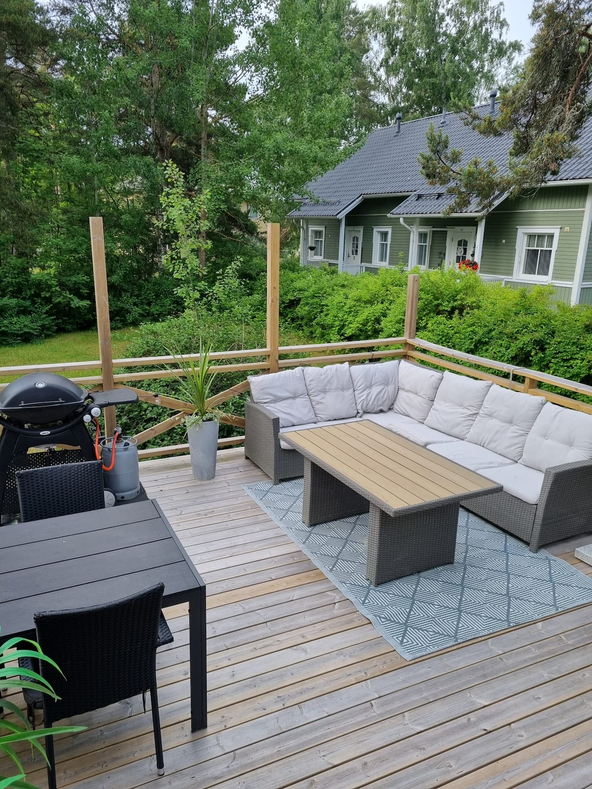 A spacious outdoor terrace features a sectional seating area with soft cushions and a wooden coffee table. A dining table with black chairs provides additional seating. Greenery surrounds the area, enhancing the feeling of privacy. A grill is positioned nearby, ready for use.