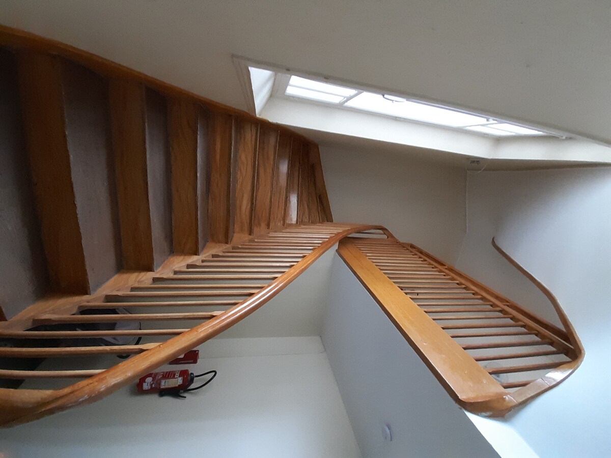 A wooden staircase features a curved design with smooth railing, leading to a well-lit upper level. Natural light filters through a skylight above, illuminating the steps and highlighting the craftsmanship of the banister. Fire safety equipment is positioned along the wall.