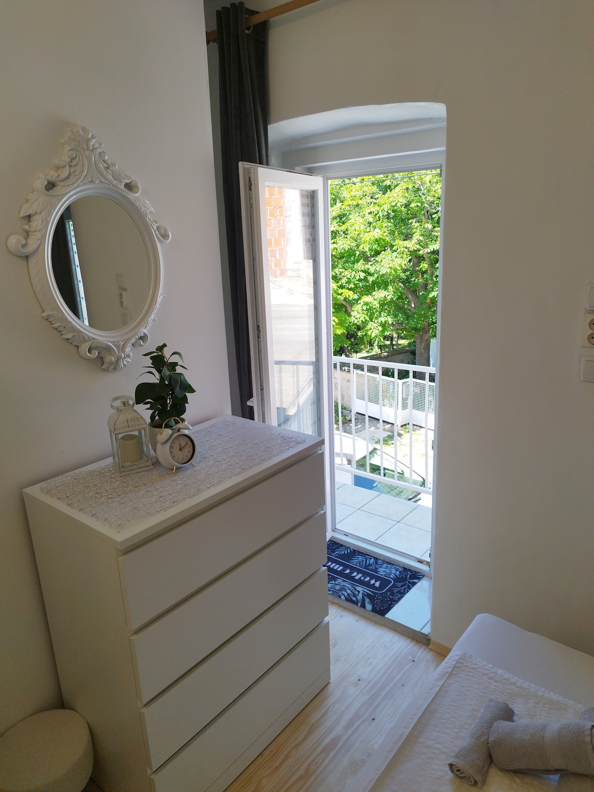 A bright room features a white dresser with four drawers, complemented by a round mirror above it. A doorway with an open balcony leads outside, allowing natural light to fill the space. A small potted plant adds a touch of greenery.