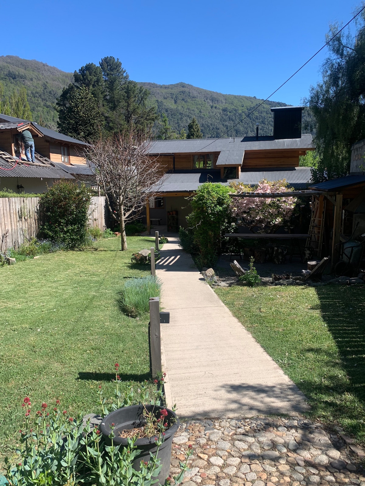 A well-maintained garden pathway is lined with lush greenery and flowering plants, leading through a tidy yard. The surrounding mountains provide a scenic backdrop, with various buildings visible in the distance under a clear blue sky.