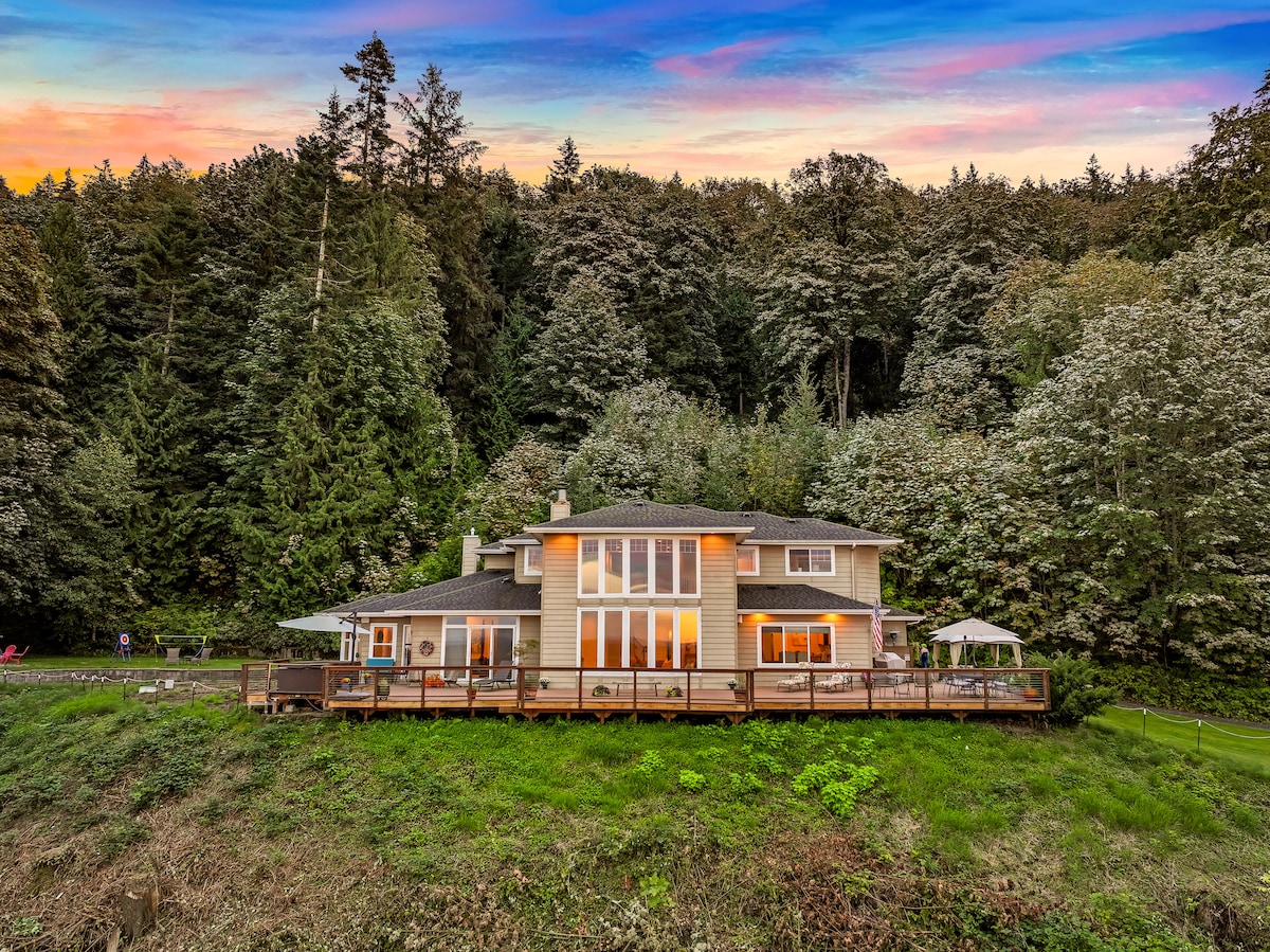 A spacious home is set against a lush backdrop of trees, featuring large windows that reflect the vibrant sunset. The expansive deck offers seating areas and is bordered by greenery, creating a serene atmosphere in a natural setting.