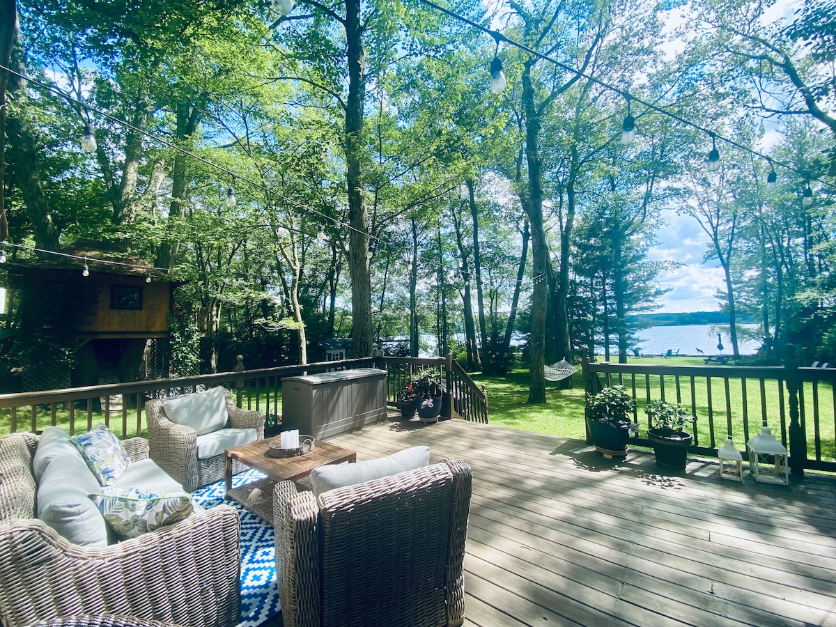 A spacious deck is surrounded by tall trees, offering a serene lake view. Comfortable seating with plush cushions is arranged around a central table. Potted plants add greenery, while string lights create a charming ambiance. A hammock is visible, nestled among the trees.