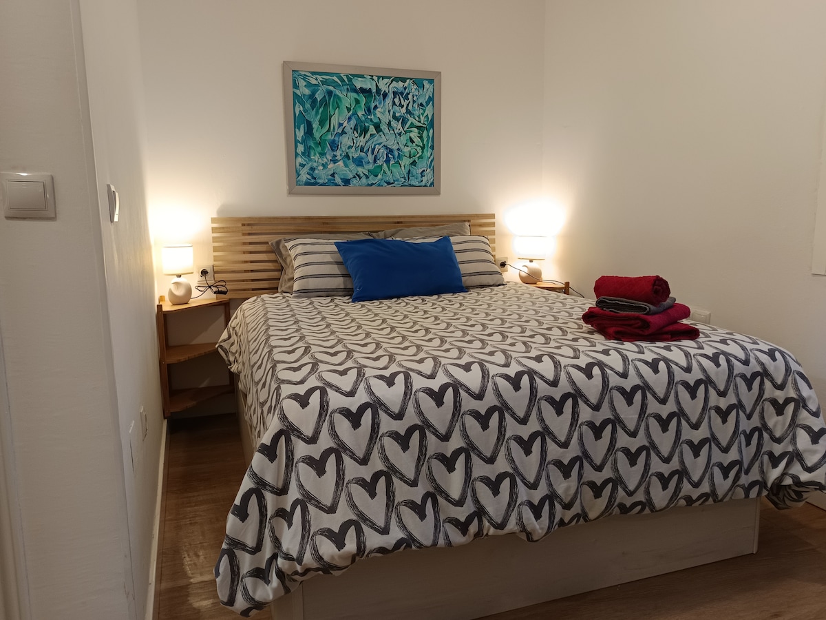 The sleeping area features a spacious bed adorned with a heart-patterned blanket and colorful throw pillows. Flanking the bed, two bedside tables hold lamps, providing gentle illumination. A piece of framed artwork hangs above the headboard, complementing the room's calming decor.