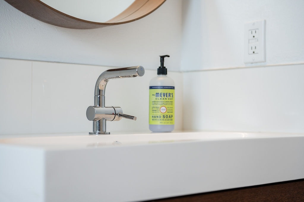 A modern bathroom sink is presented, featuring a sleek faucet and a rectangular basin. A round mirror is mounted above the sink, enhancing the space's contemporary feel. Hand soap is displayed nearby, providing a functional detail in the minimalist design.