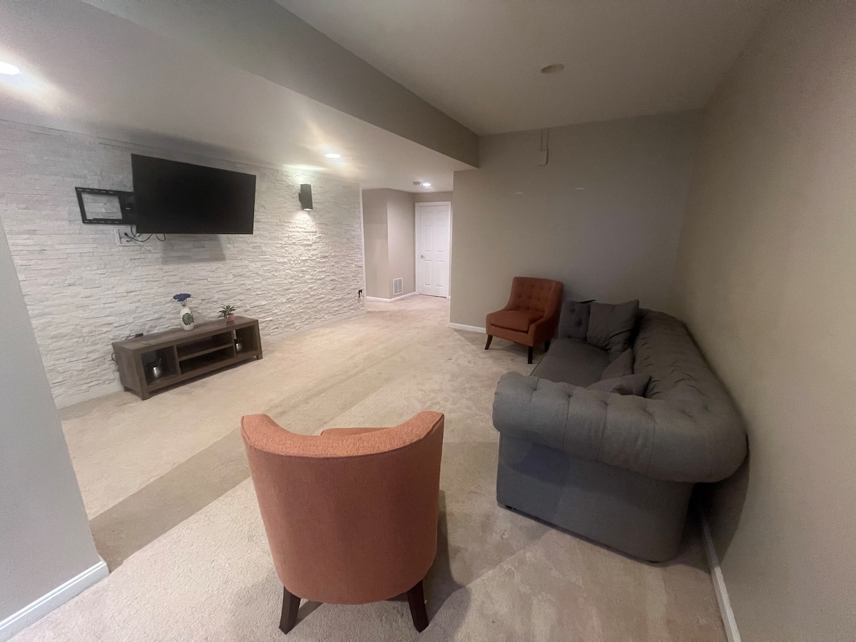 A living room area features a grey sectional sofa and an orange accent chair positioned around a low wooden table. A wall-mounted television is visible, with soft lighting illuminating the textured light-colored wall. The carpeted floor provides a comfortable atmosphere.