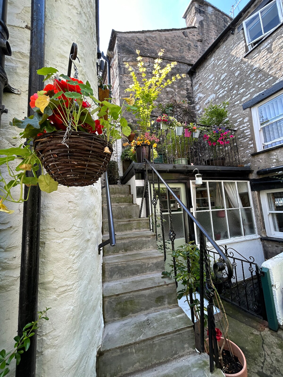 A narrow staircase leads to a quaint outdoor area, flanked by a charming stone wall. A wicker basket filled with vibrant flowers hangs beside the steps. Colorful flower pots adorn the railing, adding to the inviting atmosphere of the space.