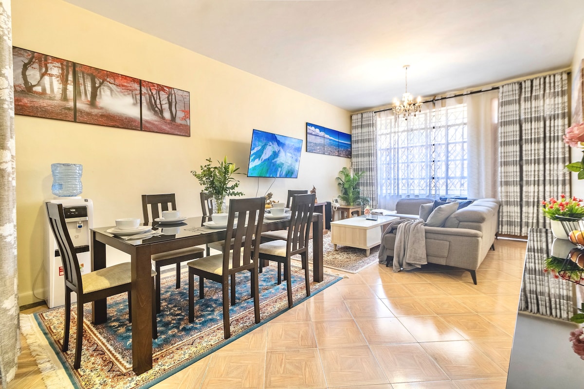 The dining area features a wooden table with four chairs, complemented by a water dispenser. A wall-mounted TV displays a scenic image. A cozy seating arrangement includes a grey sofa and coffee table, situated near large windows that allow natural light to fill the space.