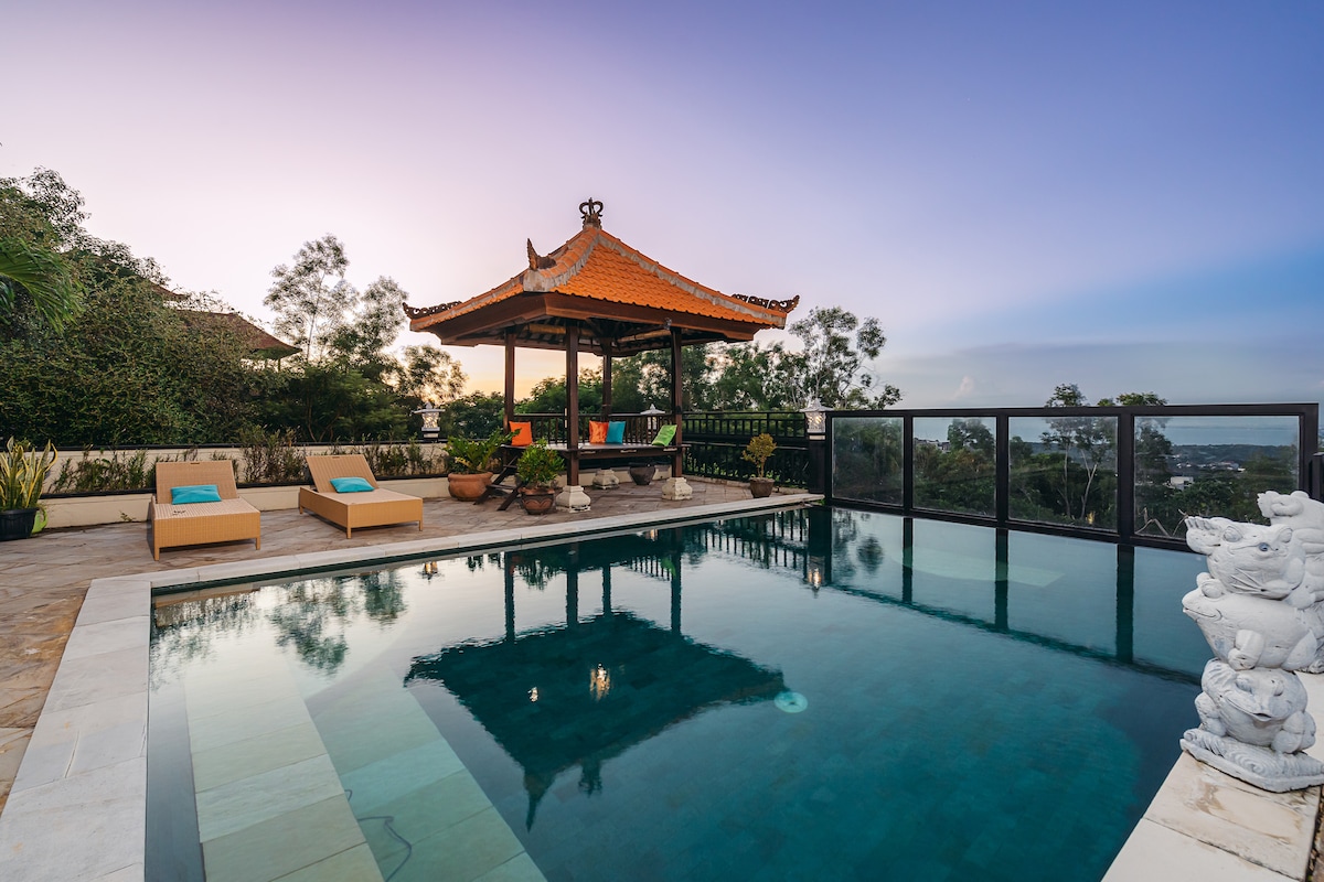 A villa pool area is featured, showcasing a serene blue pool surrounded by a stone deck. Two lounge chairs are positioned nearby under a traditional gazebo with a distinctive orange roof. Lush greenery is visible in the background, enhancing the tranquil setting.