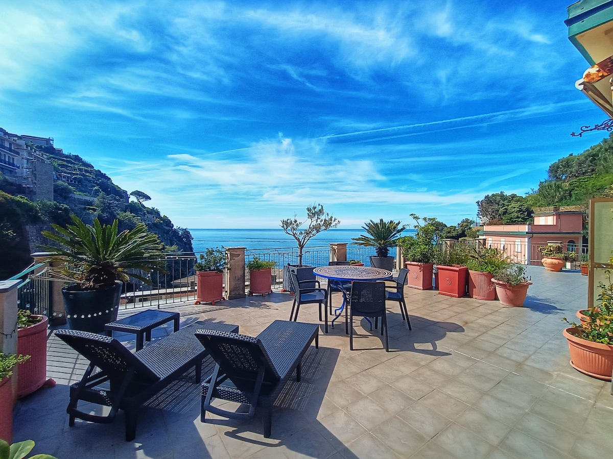 A spacious terrace is filled with comfortable lounge chairs and a round dining table surrounded by potted plants. The view extends to the serene sea and lush hills, highlighted by a clear blue sky, creating an inviting outdoor space.
