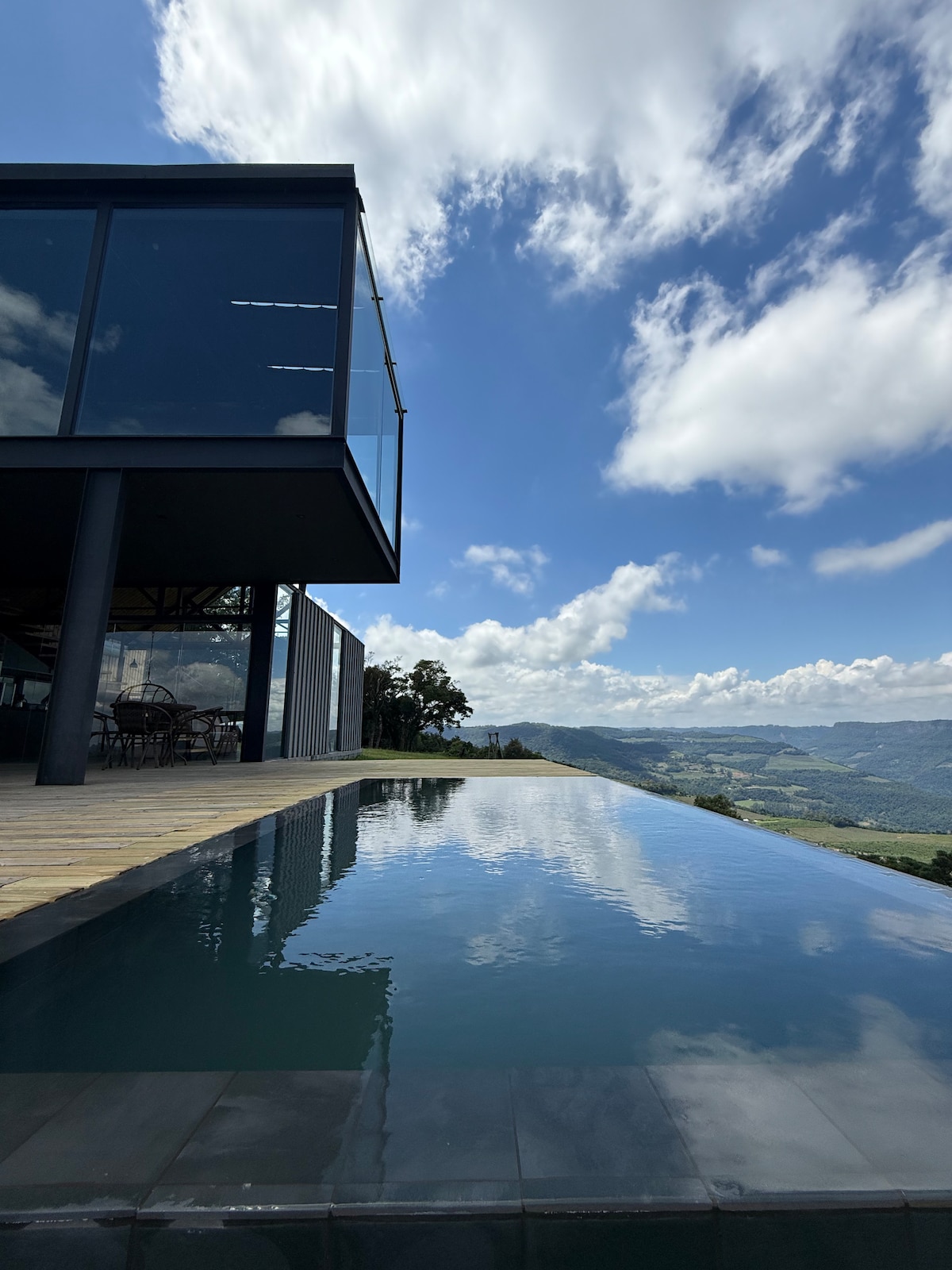 An infinity pool reflects the vibrant blue sky dotted with white clouds. The pool, surrounded by wood decking, appears to blend seamlessly with the landscape. A modern structure can be seen in the background, showcasing large glass windows that invite natural light.