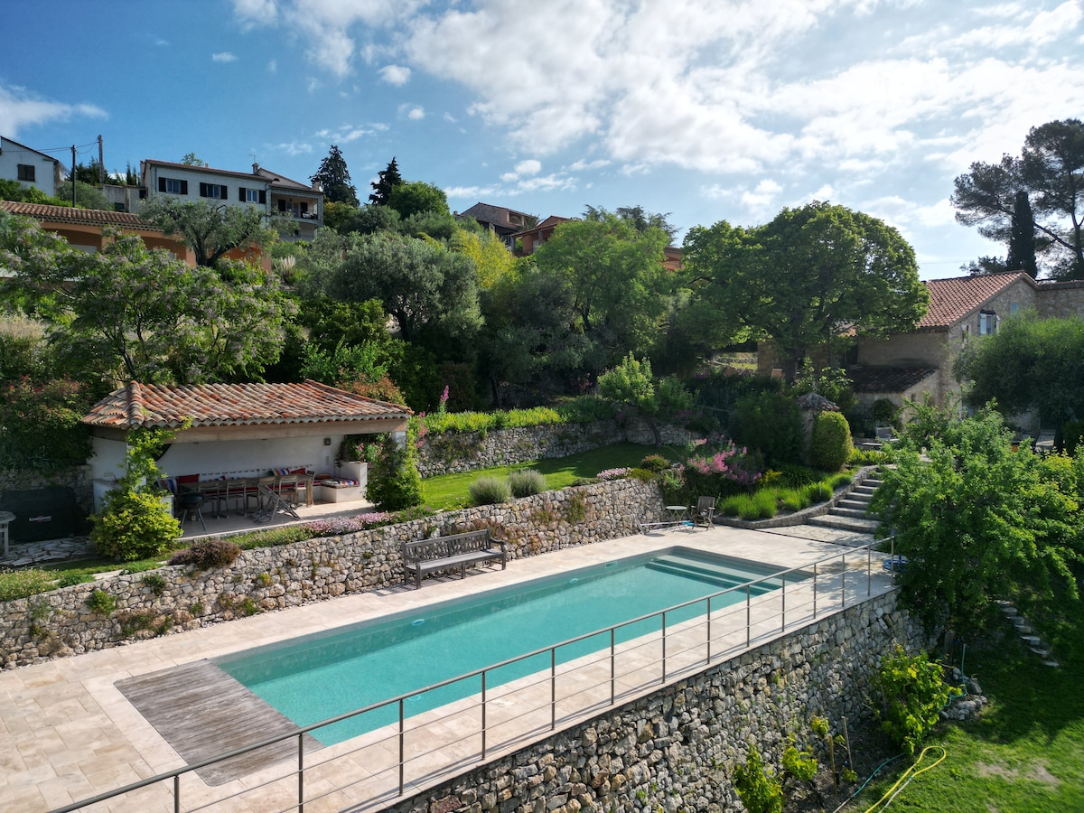A spacious swimming pool, measuring 12x4 meters, is highlighted with a surrounding stone terrace. Lush greenery and neatly arranged gardens are visible in the background, along with a covered outdoor dining area with a large table. A sunbathing area is positioned beside the pool, enhancing the serene atmosphere.