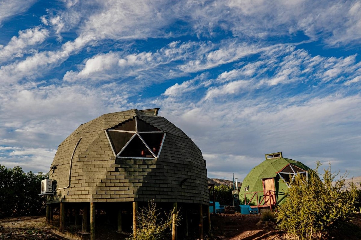 Two geodesic domes are nestled against a backdrop of blue skies with scattered clouds. Each dome features a unique green and gray exterior, with angular windows allowing light to filter in. Surrounding vegetation adds a natural element to the serene landscape.