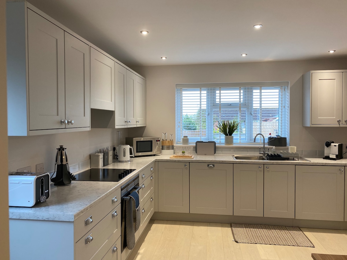 A well-equipped kitchen features modern cabinetry and a neutral color palette. Essential appliances include a microwave, kettle, and toaster, with ample counter space for food preparation. Natural light enters through a window adorned with slatted blinds, complementing the open and functional layout.