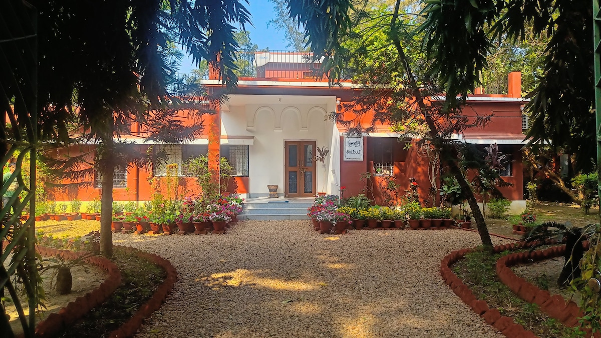 The exterior of Bulbuli is presented, featuring a charming red facade complemented by lush greenery. Flower pots are arranged along the pathway leading to the entrance, framed by soft gravel. The entrance is highlighted by a set of double doors and a welcoming porch.