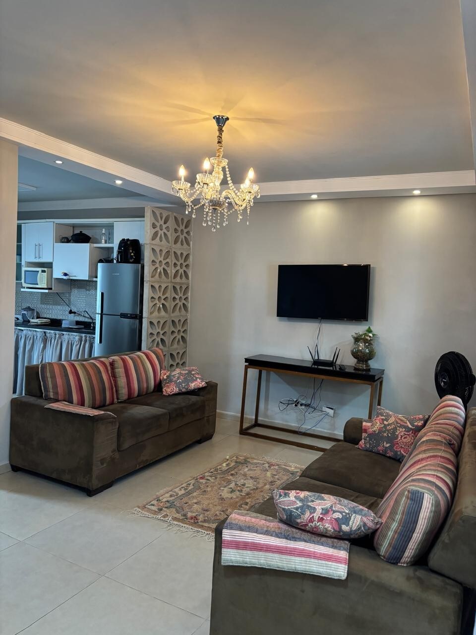 A cozy living area is presented with two brown sofas featuring patterned cushions. A decorative chandelier hangs from the ceiling, illuminating the space. A television is mounted on the wall opposite a wooden console table, and a patterned rug lies on the tiled floor.