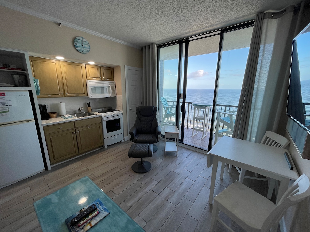 A bright living area features a compact kitchen with essential appliances, including a microwave and a glass stovetop oven. A comfortable chair is positioned near a window, offering a view of the ocean. Light-colored tiles reflect natural light from the large sliding door that leads to the balcony.