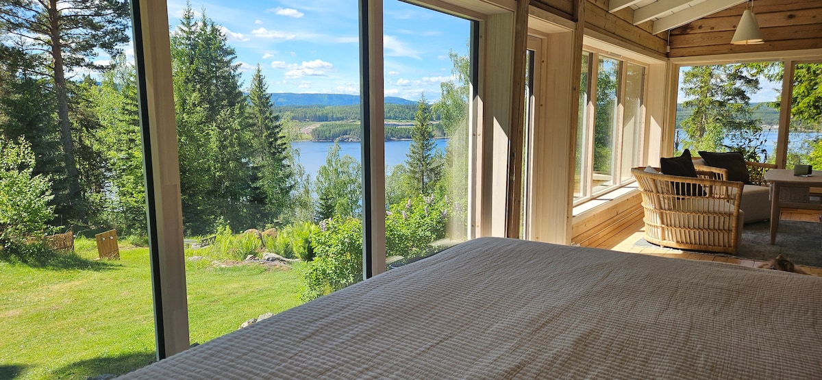 The room features large windows offering expansive views of the lush landscape and lake. A spacious bed is visible, inviting relaxation. Natural light floods the interior, enhancing the connection to the serene outdoors, surrounded by trees and greenery.